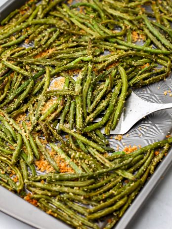 Roasted green beans from frozen on a sheet pan being scooped up with a metal spatula.