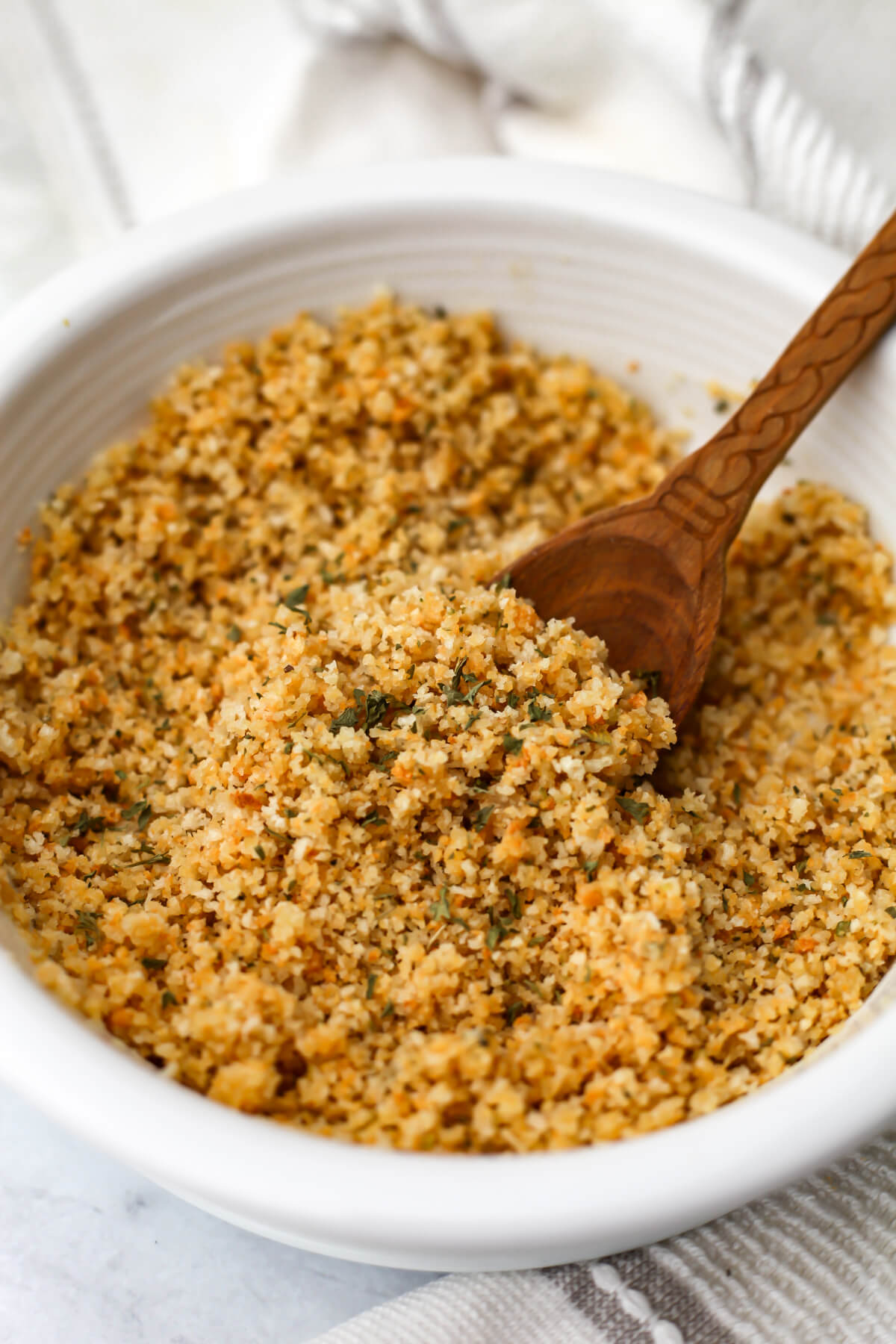 Vegan seasoned breadcrumbs in a white bowl are being stirred with a small wooden spoon.