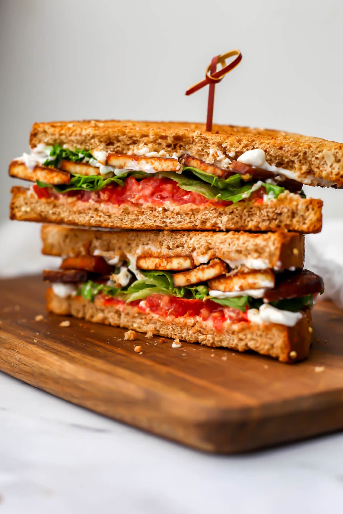 A vegan tofu BLT cut in half and stacked on top of each other on a wooden cutting board.