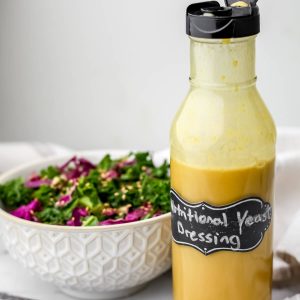 A glass bottle filled with nutritional yeast dressing sitting next to a kale salad.