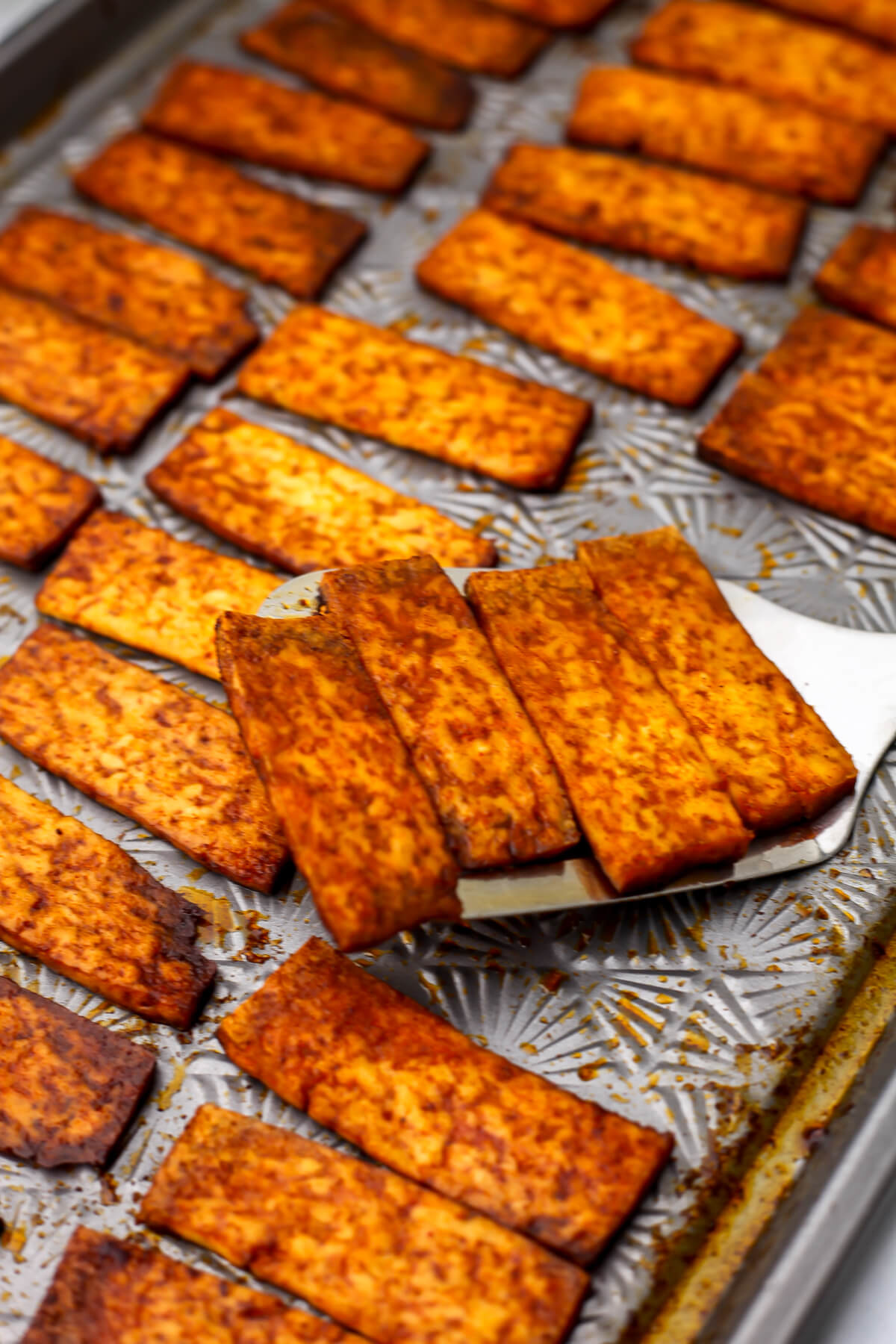 Smoky tofu bacon being picked up with a spatula off a baking sheet.