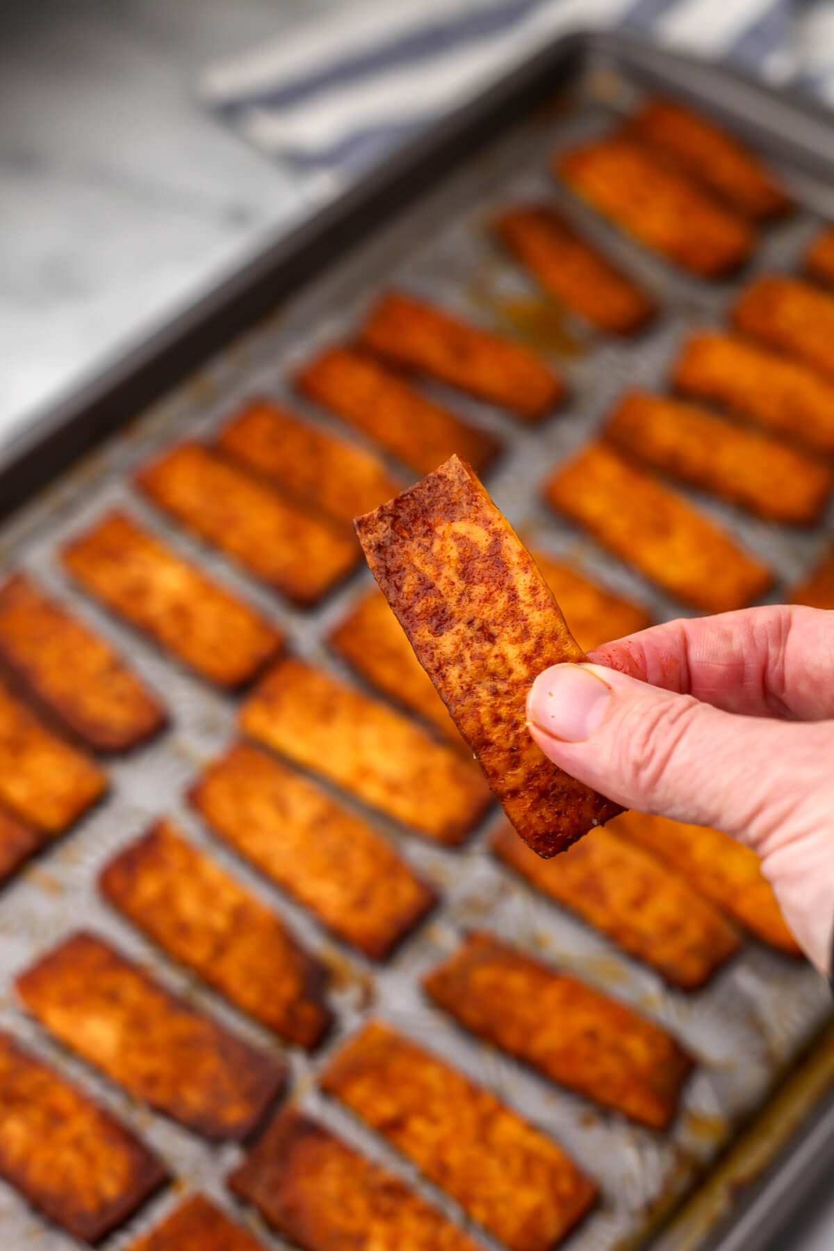 Someone holding up a piece of vegan tofu bacon above a sheet pan full of it.