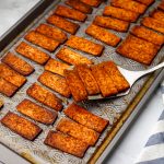 A sheet pan full of vegan smoky tofu bacon strips being picked up with a spatula.