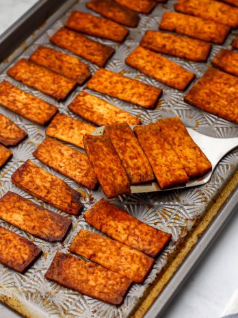 A sheet pan full of vegan smoky tofu bacon strips being picked up with a spatula.