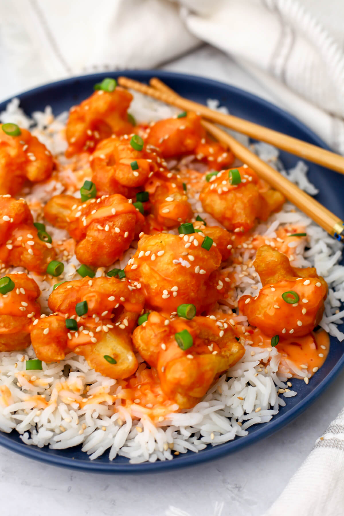 A blue plate with white rice, deep-fried cauliflower and bang bang sauce drizzled over it. Garnished with green onions and sesame seeds.