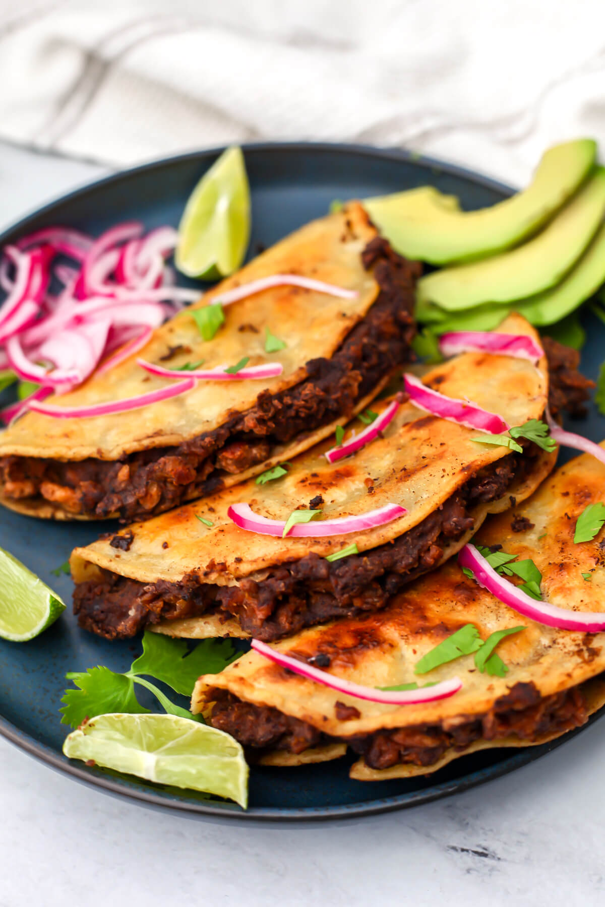 Three crispy black bean tacos with pickled red onions and avocado on the side.