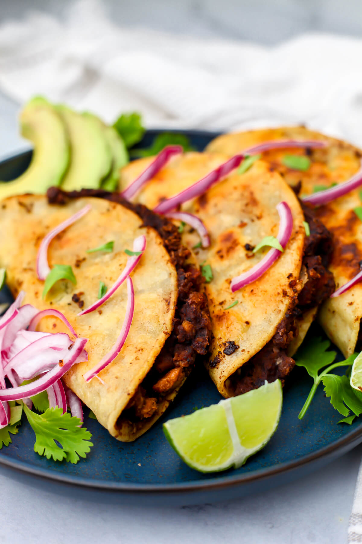 Three vegan black bean tacos on a blue plate with Mexican pickled onions and sliced avocado on the side.