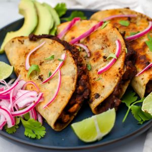Three vegan black bean tacos on a blue plate with pickled red onions, cilantro, and lime wedges.