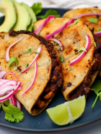 Three vegan black bean tacos on a blue plate with pickled red onions, cilantro, and lime wedges.