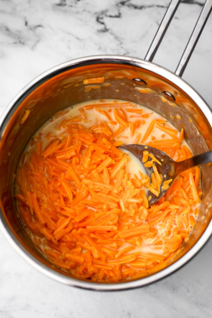 Vegan shredded cheese and plant milk in a sauce pan being stirred with a large spoon.