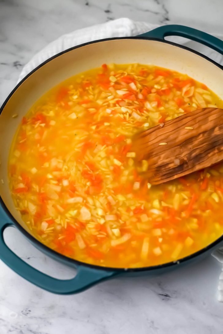 Vegan chicken broth added to the onions, carrots, and orzo in a skillet.