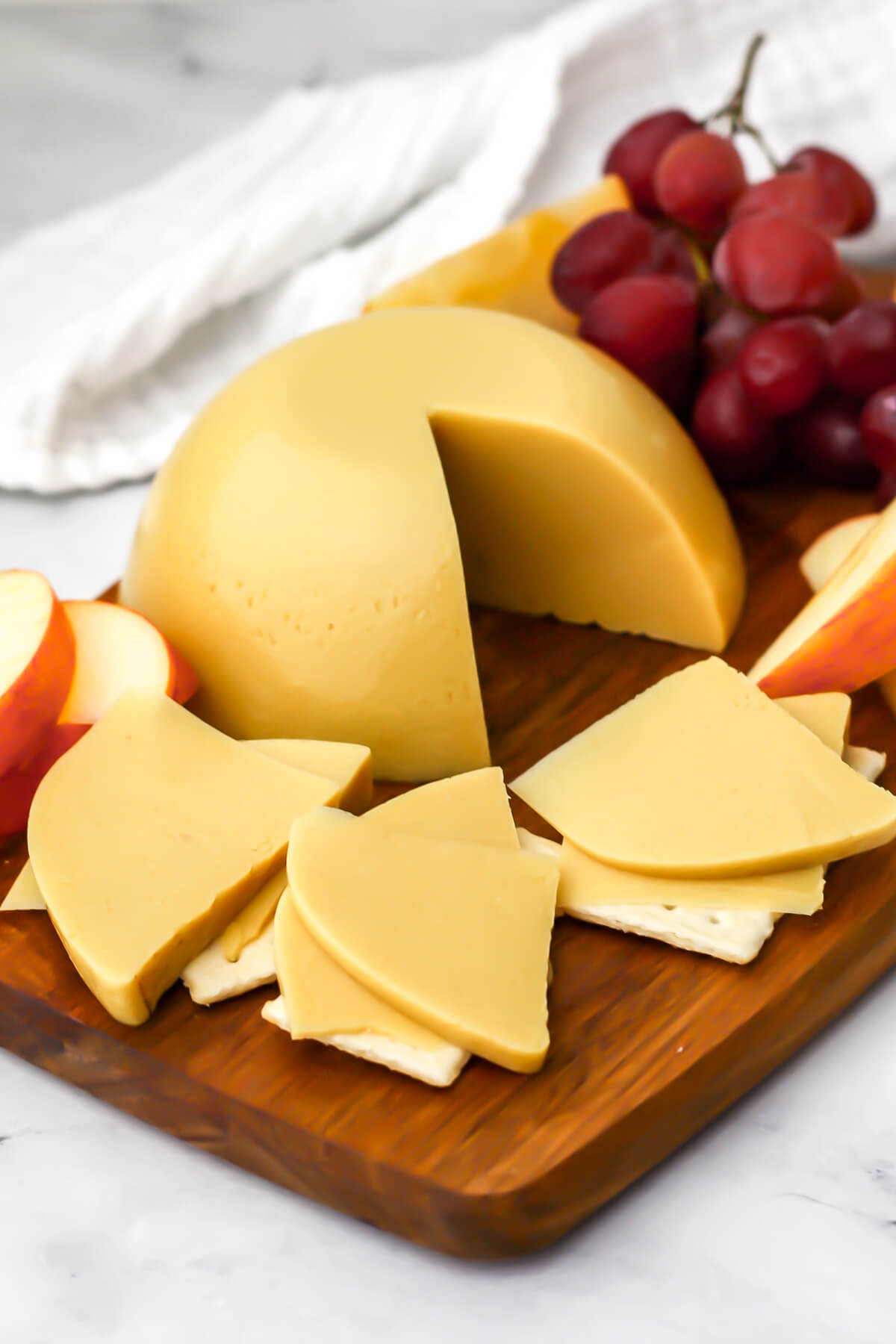 A wooden cutting board with sliced oat milk cheese layered on crackers with grapes and apple slices on the side.