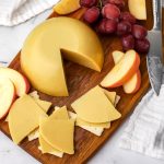 A top view of a block of oat milk cheese cut into slices and served on crackers with grapes and apples on the side.