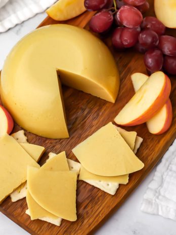 A top view of a block of oat milk cheese cut into slices and served on crackers with grapes and apples on the side.